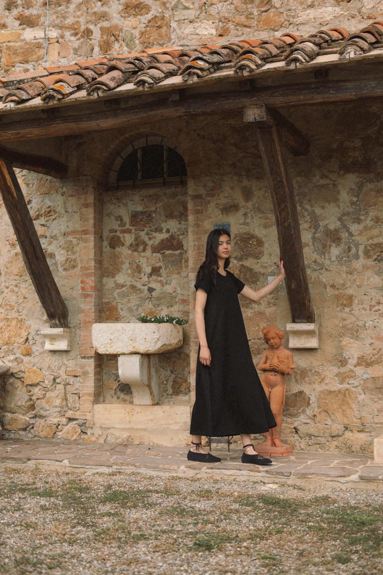 Model wearing the Palmer Dress in Noir by Gillian Stevens, full-length side view at a rustic Tuscan stone building