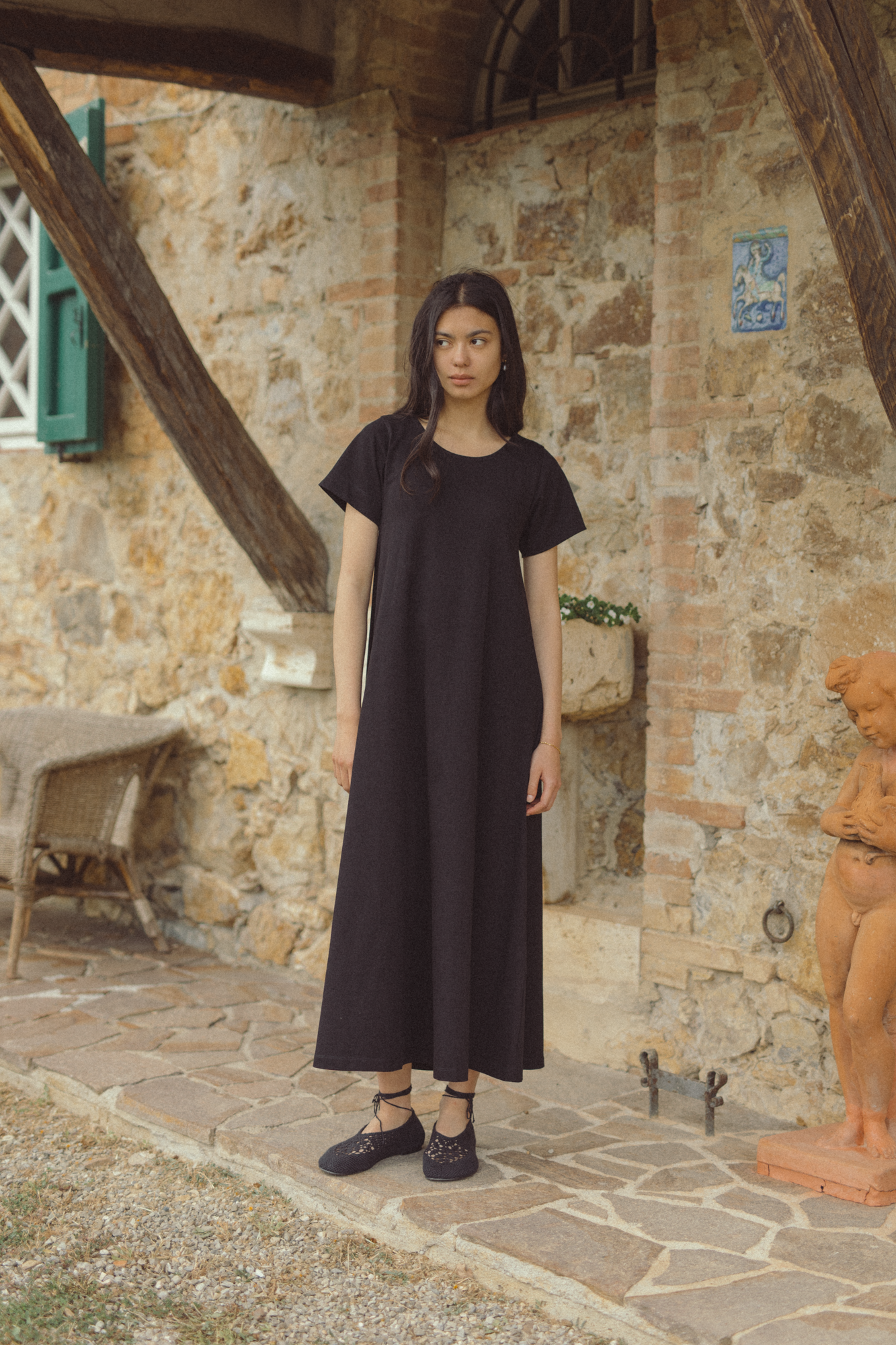 Model wearing the Palmer Dress in Noir by Gillian Stevens, front view against a rustic stone building