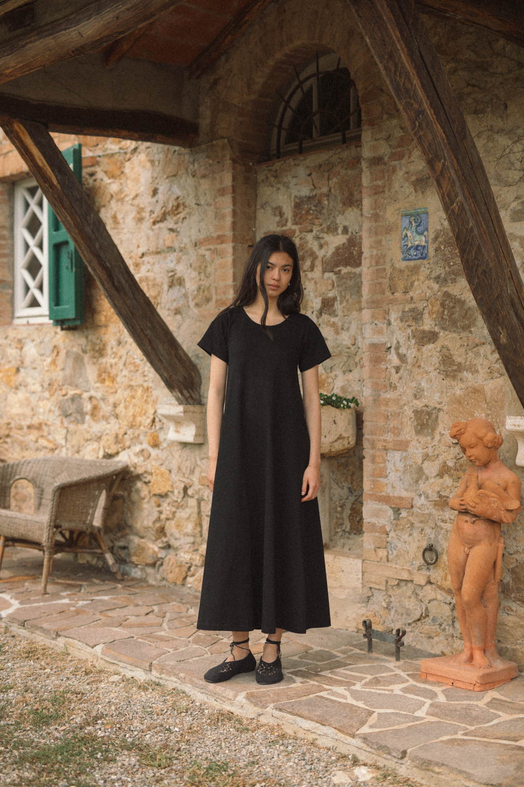 Model wearing the Palmer Dress in Noir by Gillian Stevens, front view at a rustic stone courtyard
