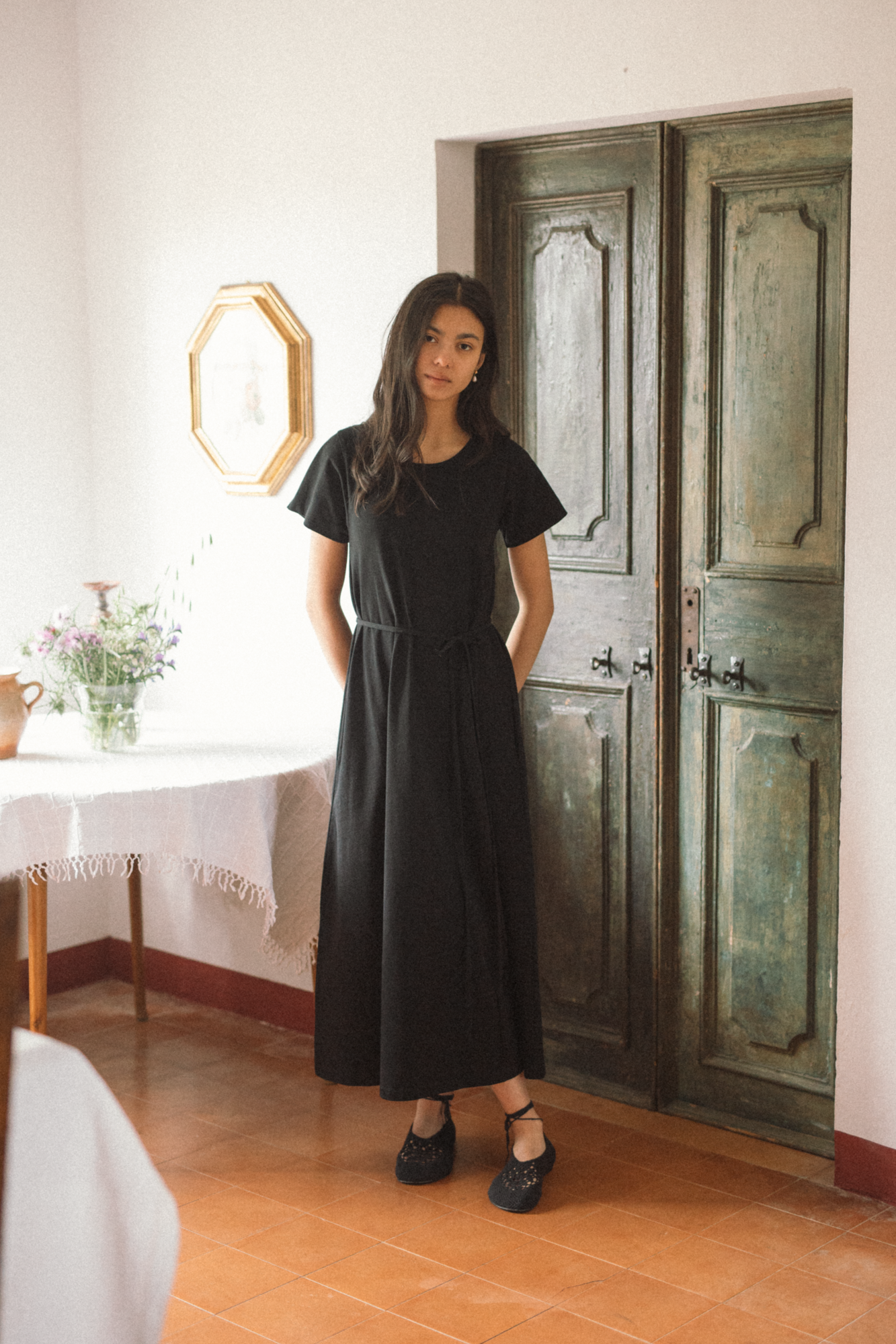 Model wearing the Palmer Dress in Noir by Gillian Stevens, front view beside a weathered green door