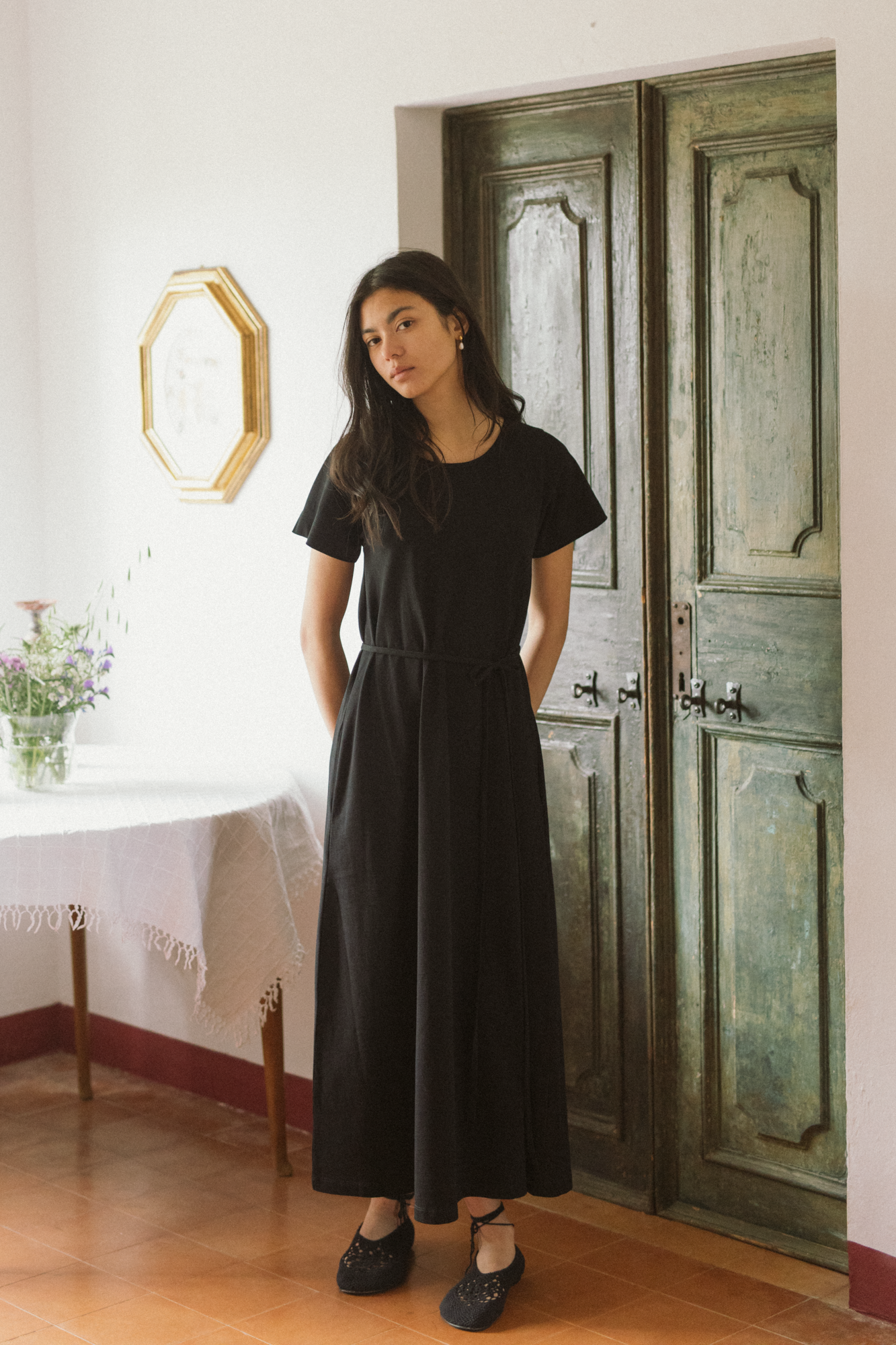 Model wearing the Palmer Dress in Noir by Gillian Stevens, front view beside a weathered green door