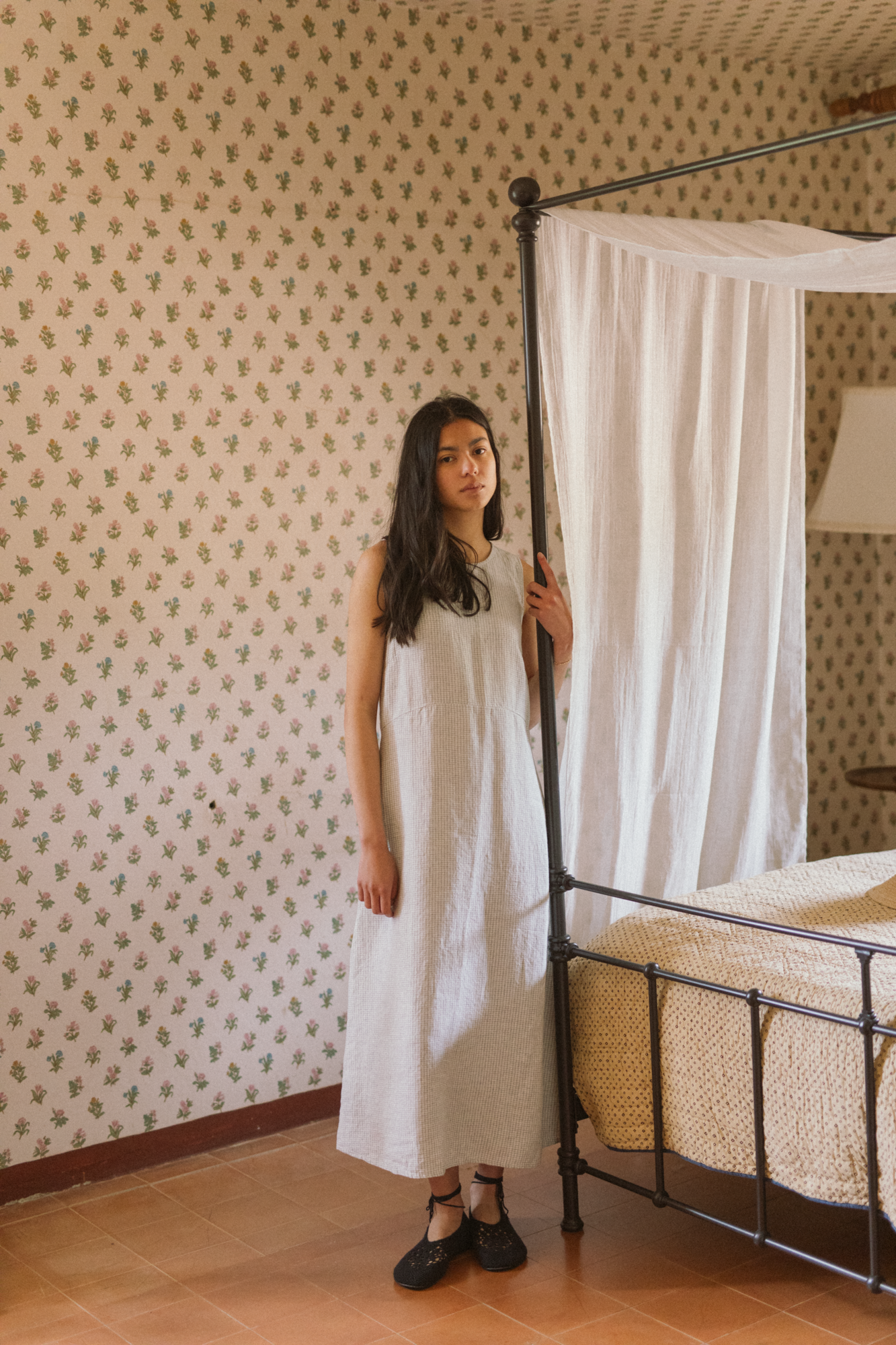 Model wearing the Paige Dress in Dusty Blue Plaid by Gillian Stevens, front view beside a four-poster bed