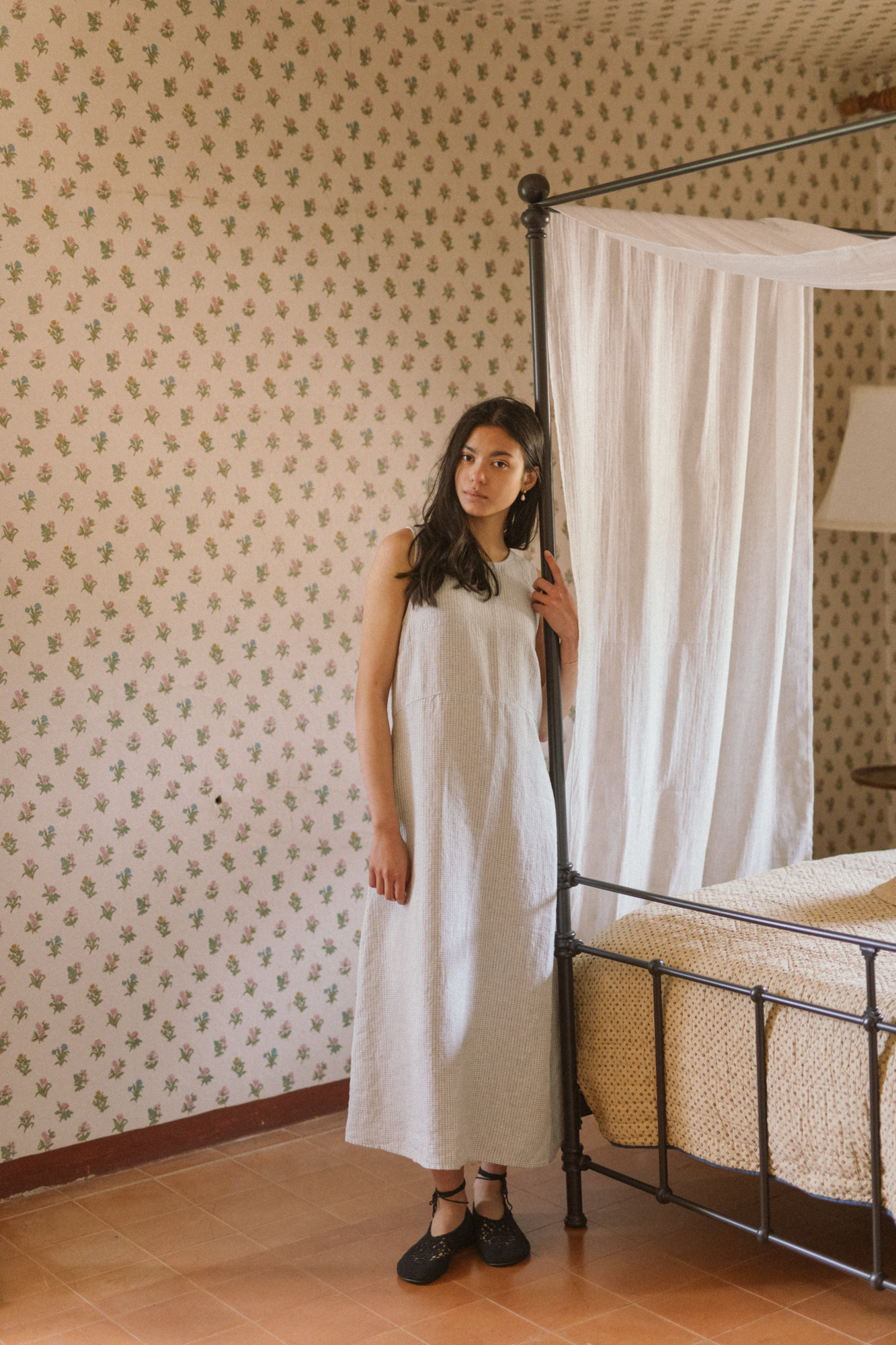 Model wearing the Paige Dress in Dusty Blue Plaid by Gillian Stevens, front view in a floral wallpapered bedroom
