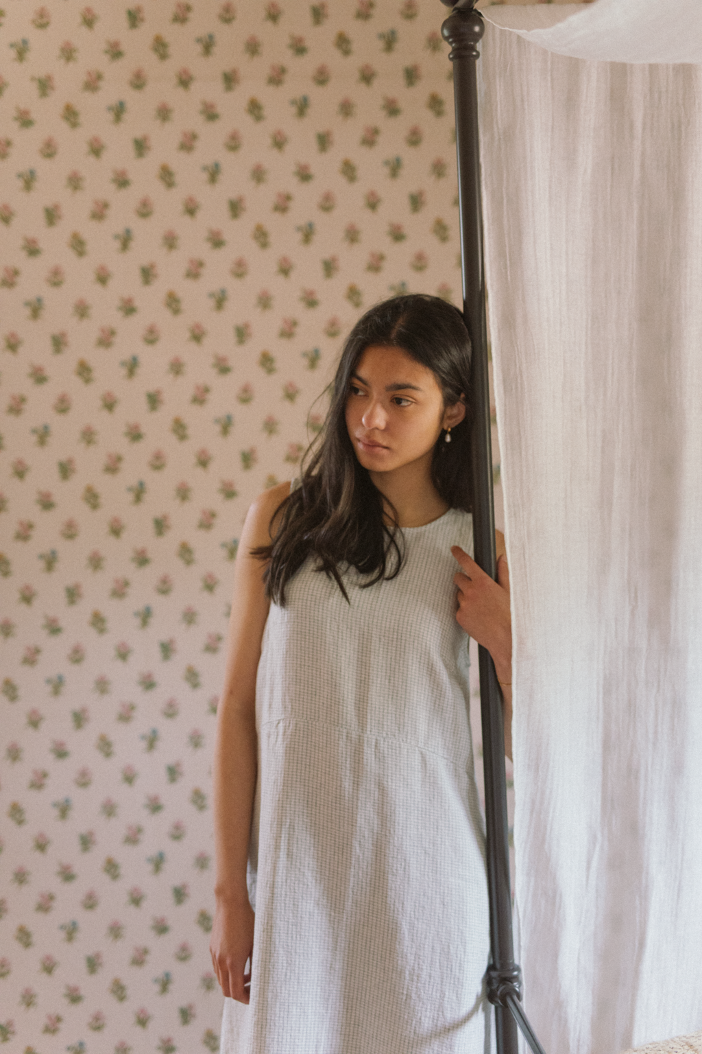 Model wearing the Paige Dress in Dusty Blue Plaid by Gillian Stevens, front view beside a four-poster bed
