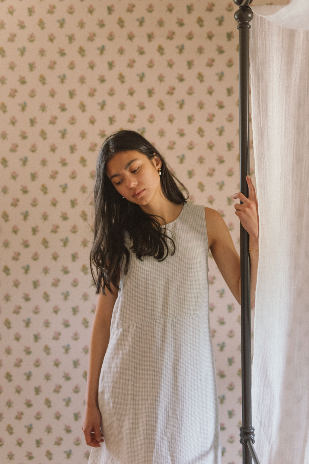Model wearing the Paige Dress in Dusty Blue Plaid by Gillian Stevens, front view beside a canopy bed post