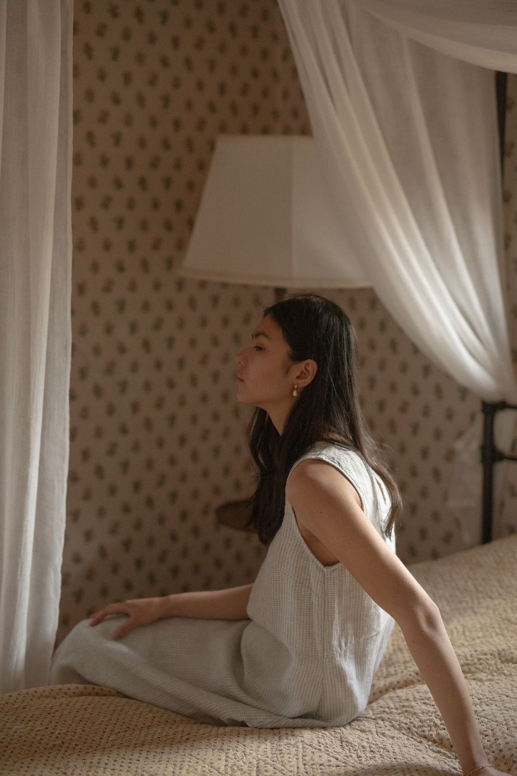 Model seated on a canopy bed in the Paige Dress in Dusty Blue Plaid by Gillian Stevens, side profile view