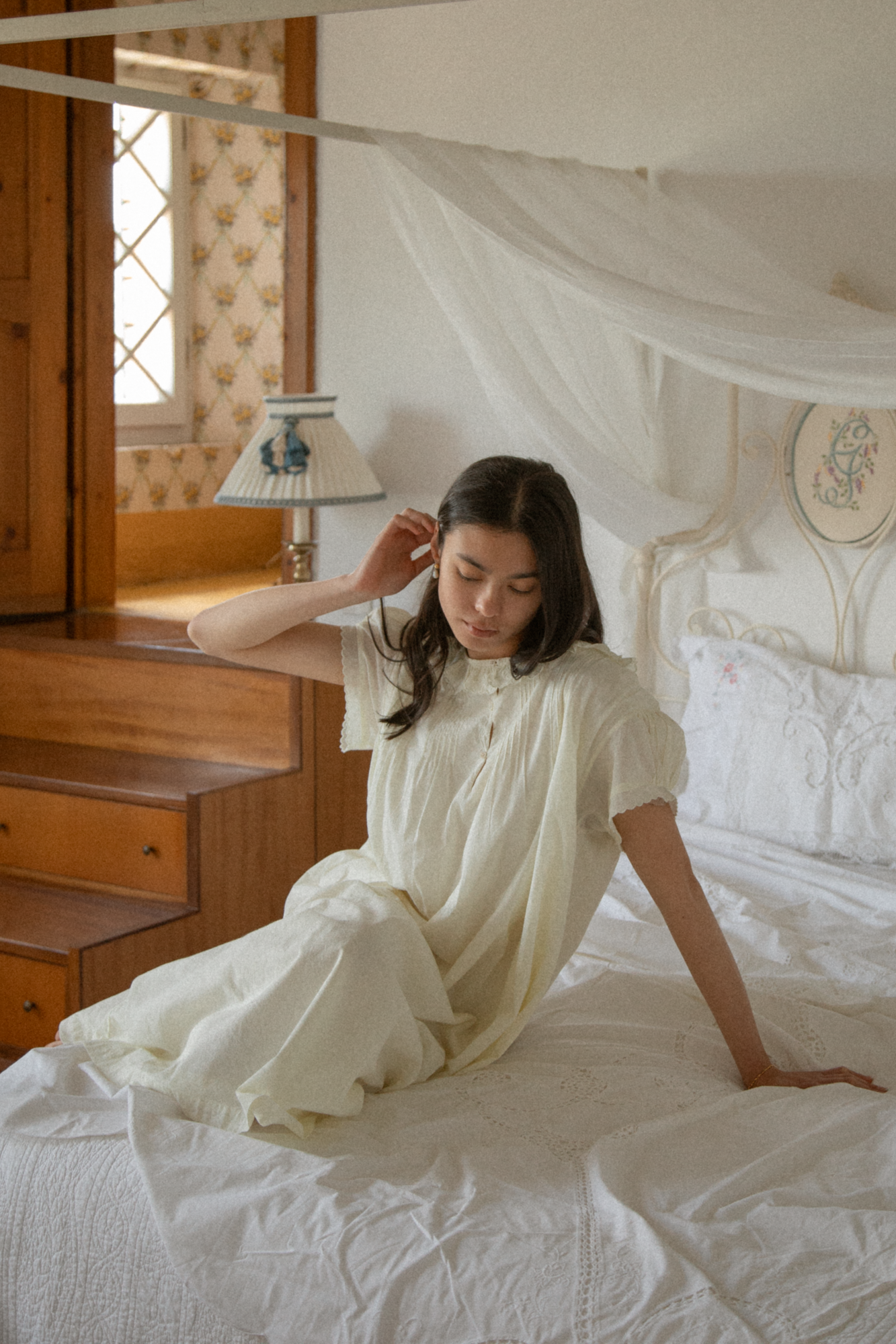 Model wearing the Clover Dress in Butter by Gillian Stevens, seated on a canopy bed, front view