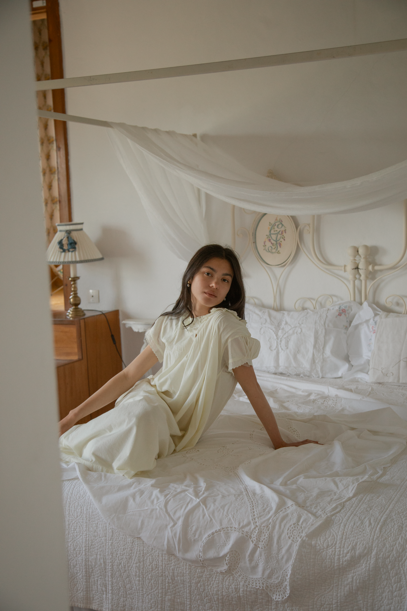 Model seated on a canopy bed wearing the Clover Dress in Butter by Gillian Stevens, full-length front view