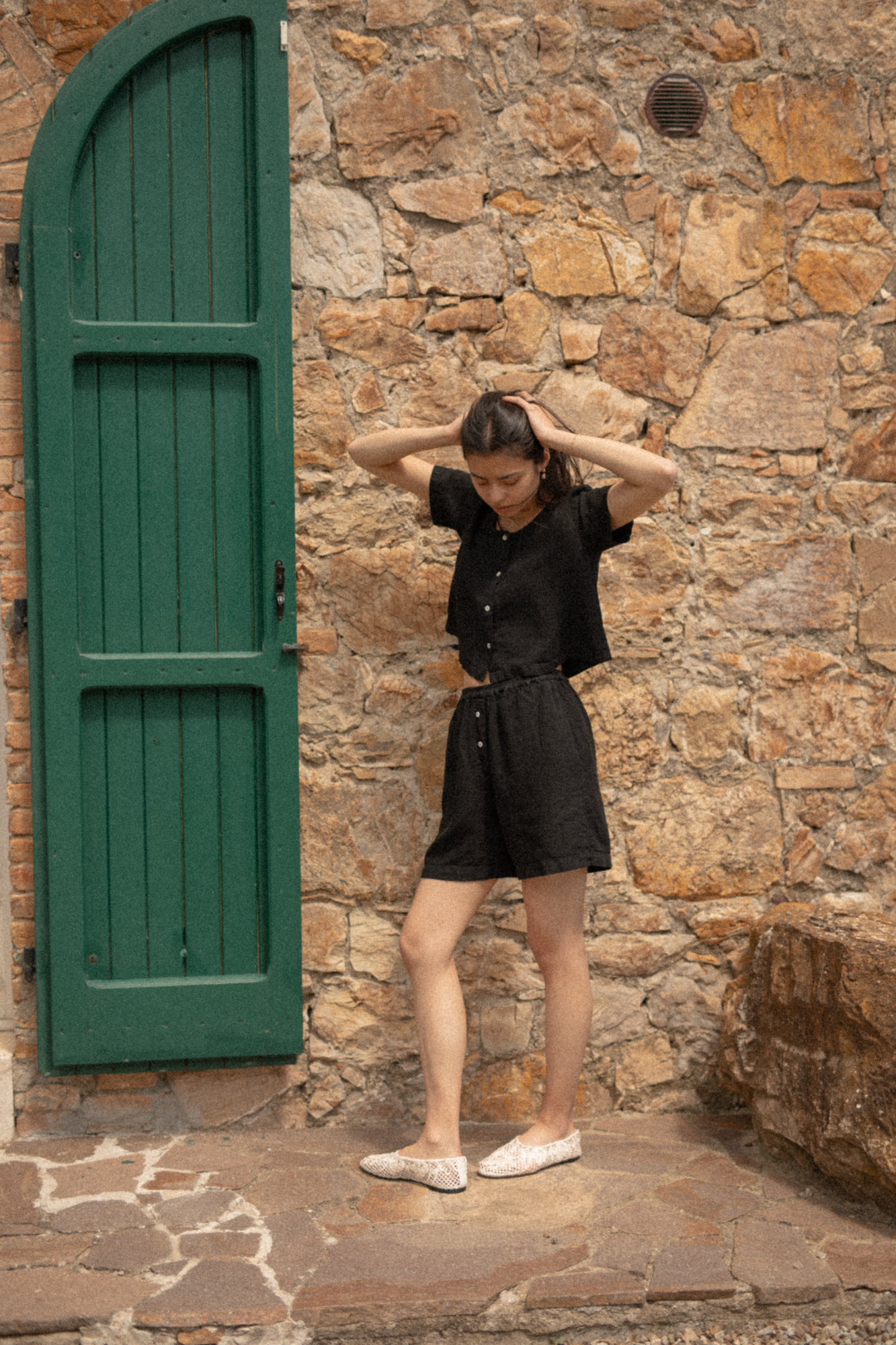 Model wearing the Wren Top in Noir by Gillian Stevens, front view beside a rustic stone wall and green door