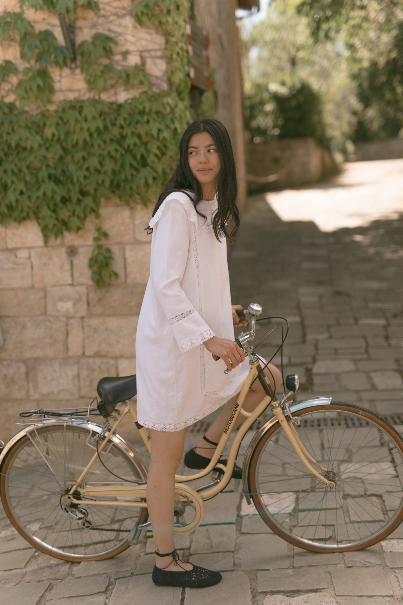 Model in the Laine Dress in Salt by Gillian Stevens, side view beside a vintage bicycle on cobblestones