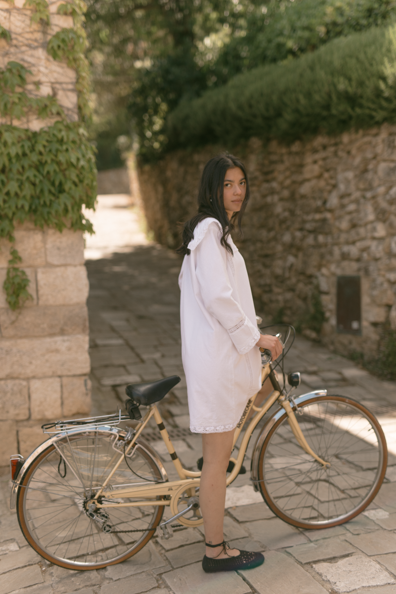 Model in the Laine Dress in Salt by Gillian Stevens, side view holding a vintage bicycle on a cobblestone lane