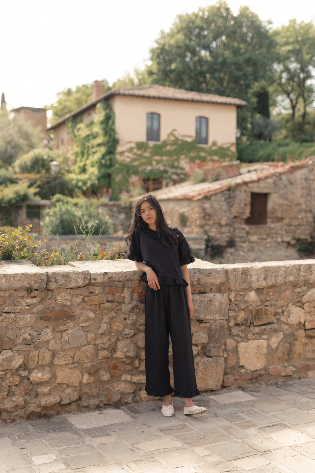 Model wearing the Arlette Pant in Noir by Gillian Stevens, full-length front view against a Tuscan stone wall