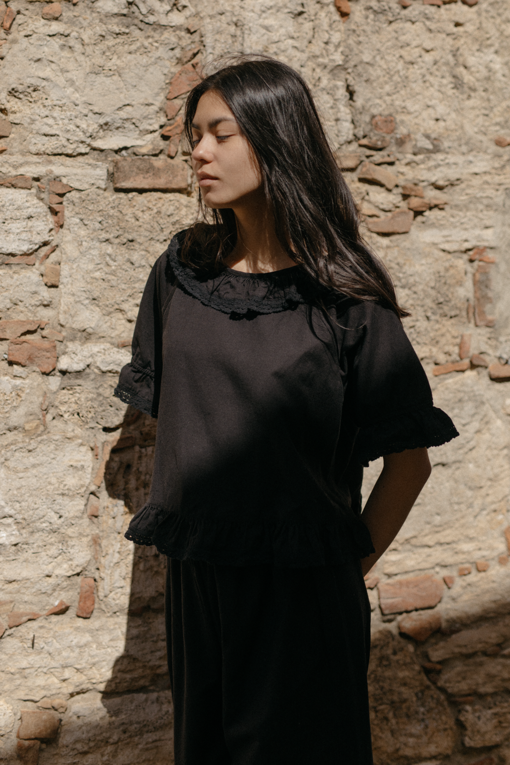 Model wearing the Arlette Top in Noir by Gillian Stevens, front view against a rustic stone wall