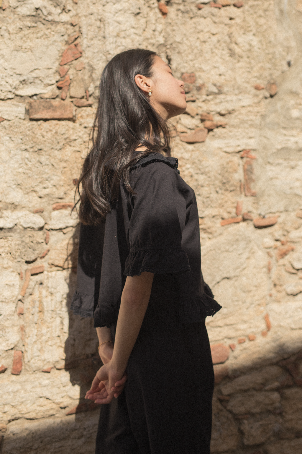 Model wearing the Arlette Top in Noir by Gillian Stevens, side view against a rustic stone wall