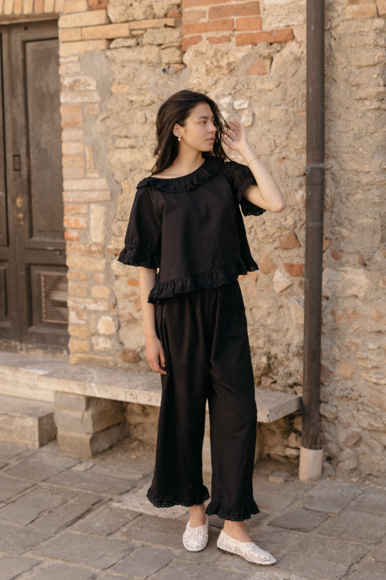 Model wearing the Arlette Top in Noir by Gillian Stevens, styled with matching black wide-leg pants, outdoor stone setting