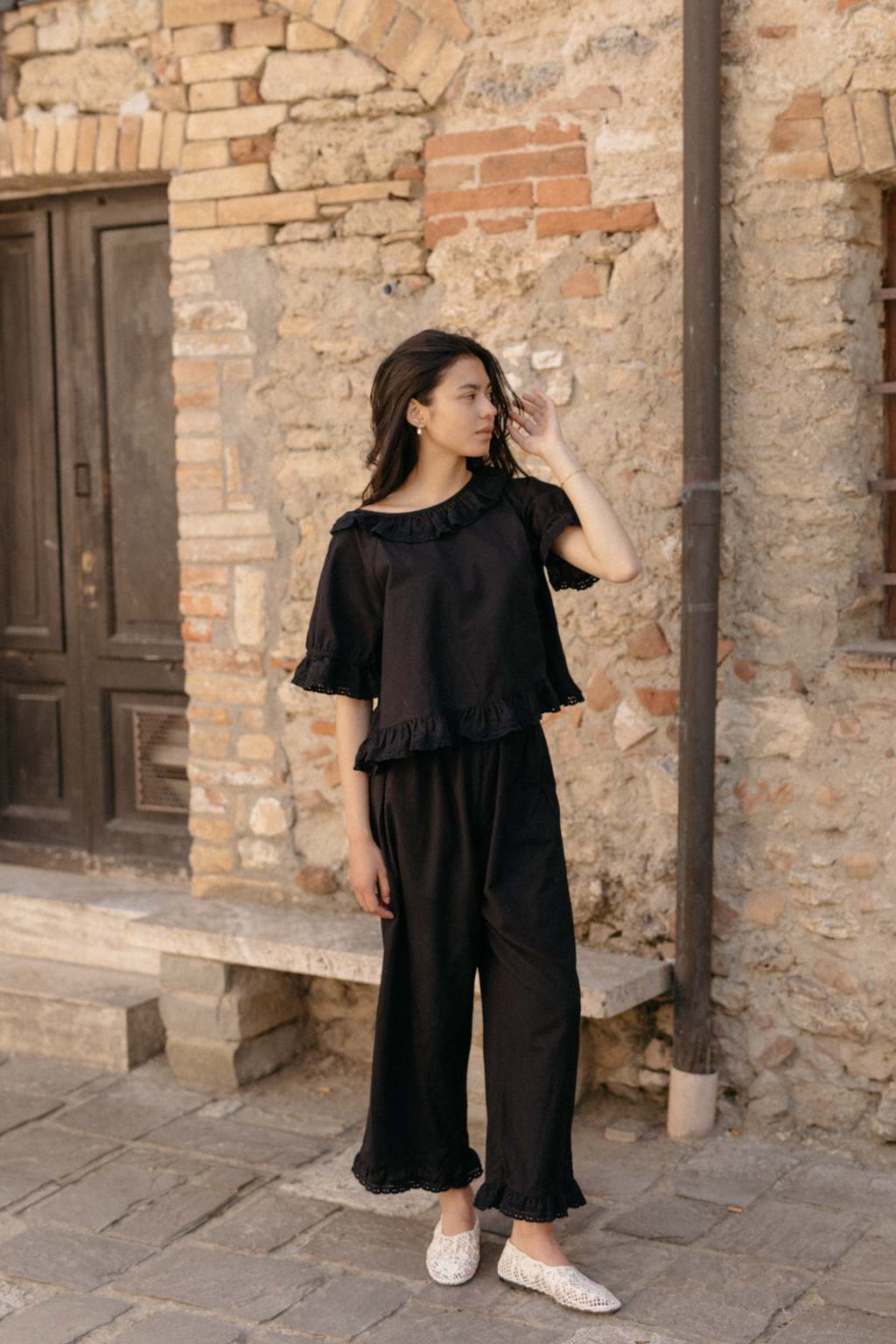 Model wearing the Arlette Pant in Noir by Gillian Stevens, full-length front view against a rustic stone wall