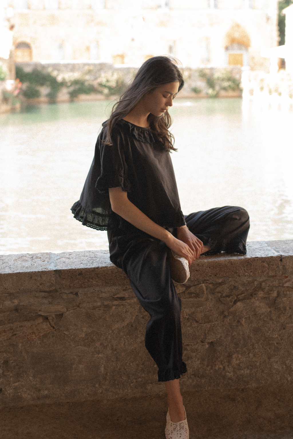 Model seated on a stone wall in the Arlette Top in Noir by Gillian Stevens, styled with matching dark trousers