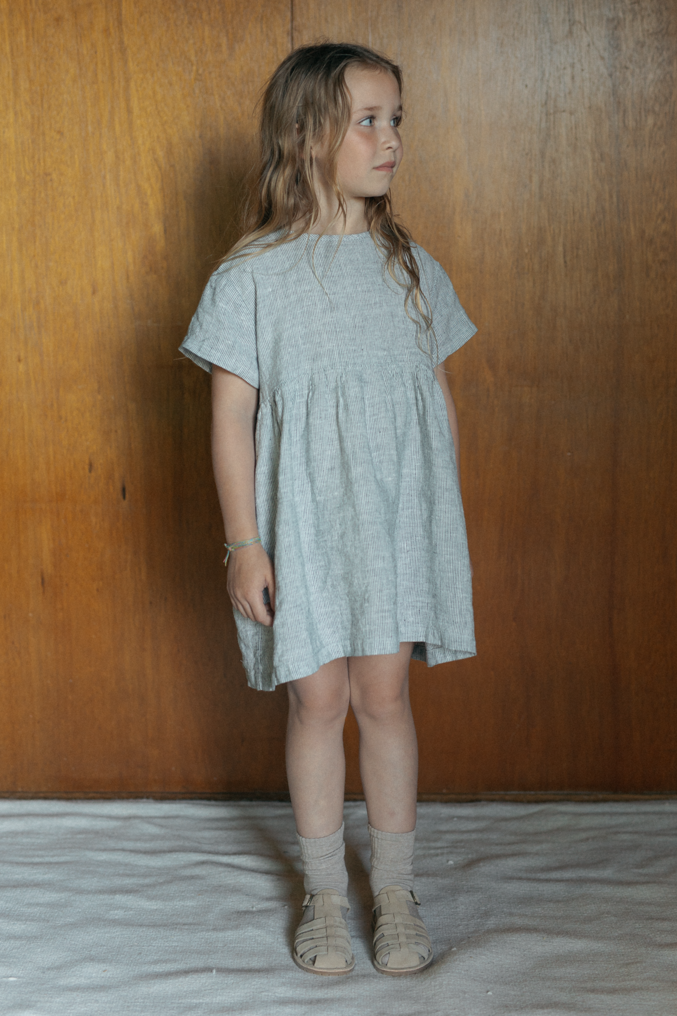 Child wearing the Mini Aya in Charcoal Stripe by Gillian Stevens, three-quarter view against warm timber panelling