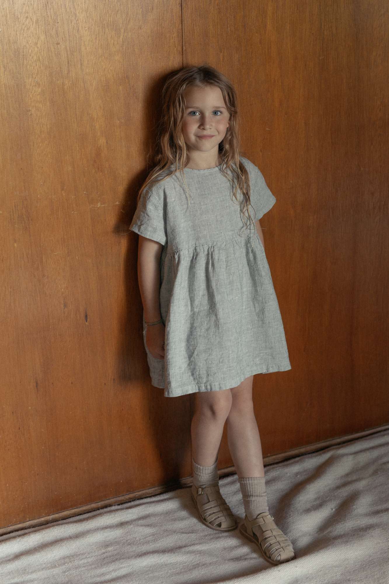 Child wearing the Mini Aya in Charcoal Stripe by Gillian Stevens, front view against warm timber wall
