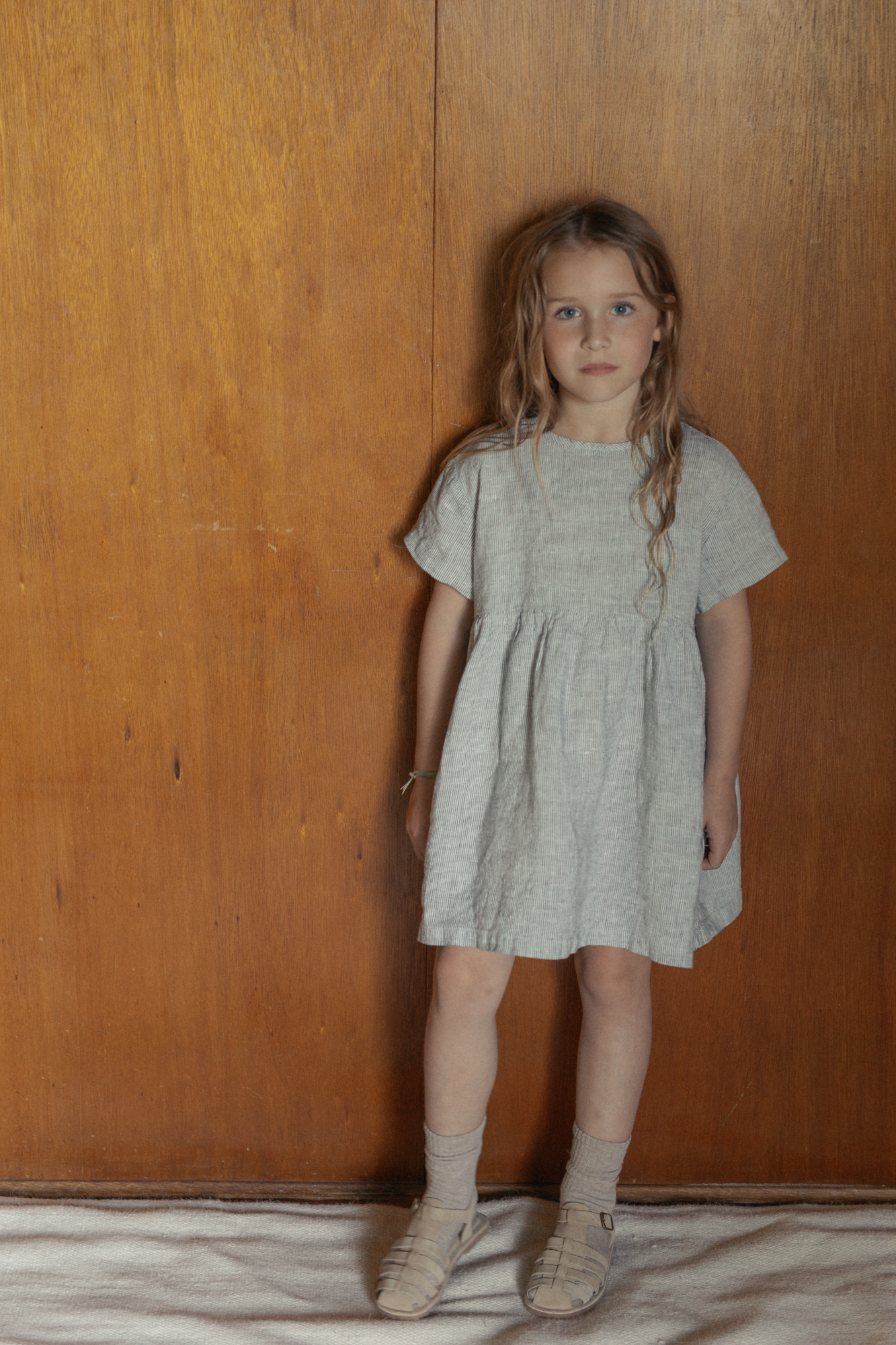 Child wearing the Mini Aya in Charcoal Stripe by Gillian Stevens, front view against a warm timber background