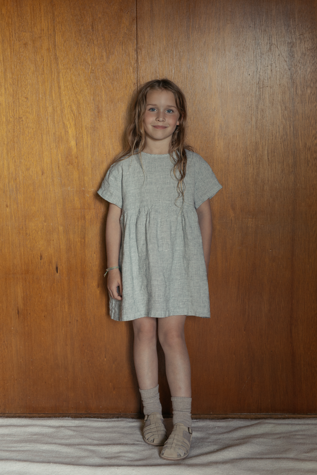 Child wearing the Mini Aya in Charcoal Stripe by Gillian Stevens, front view against a warm timber background