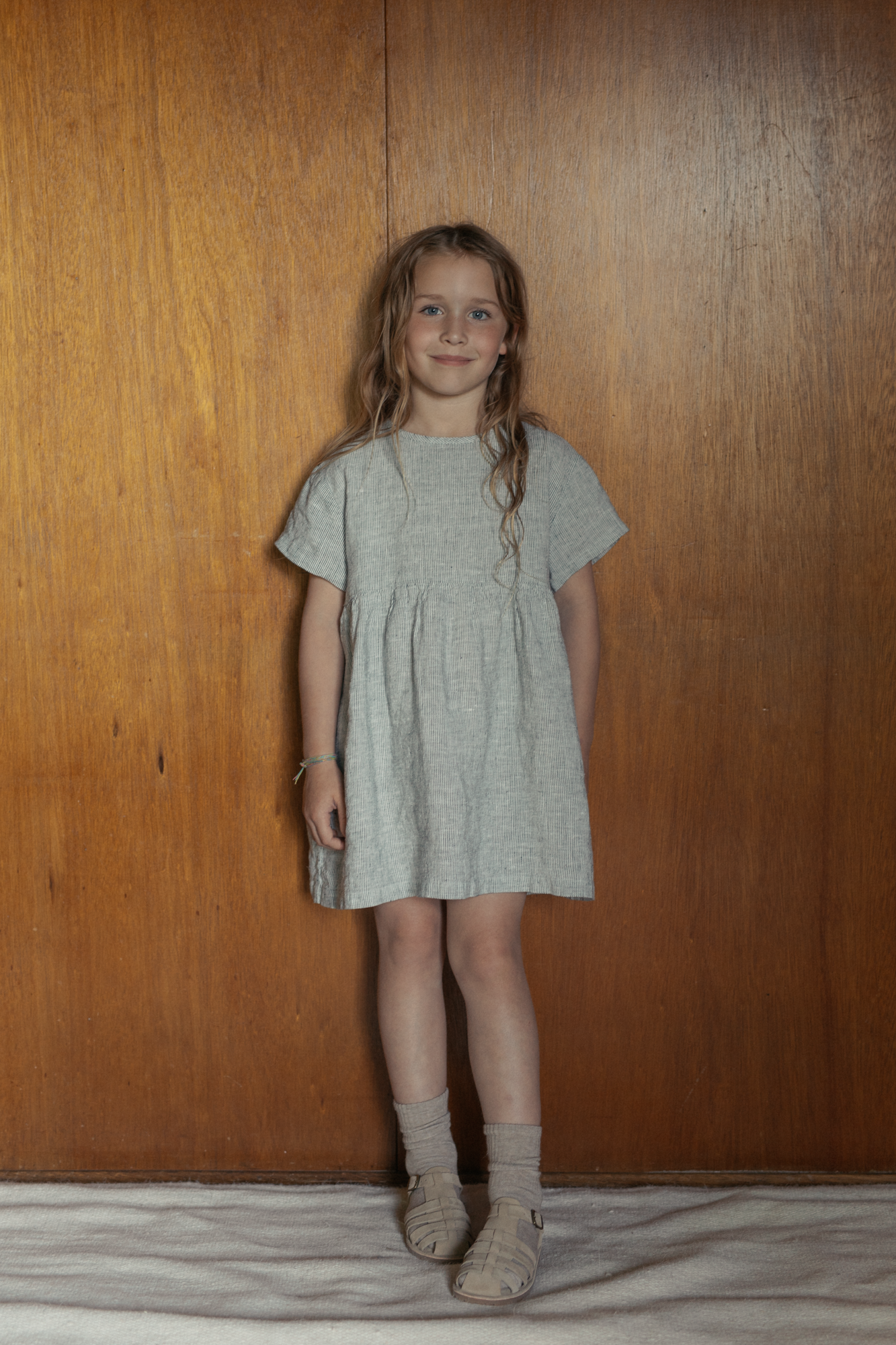 Child wearing the Mini Aya in Charcoal Stripe by Gillian Stevens, front view against a warm timber background