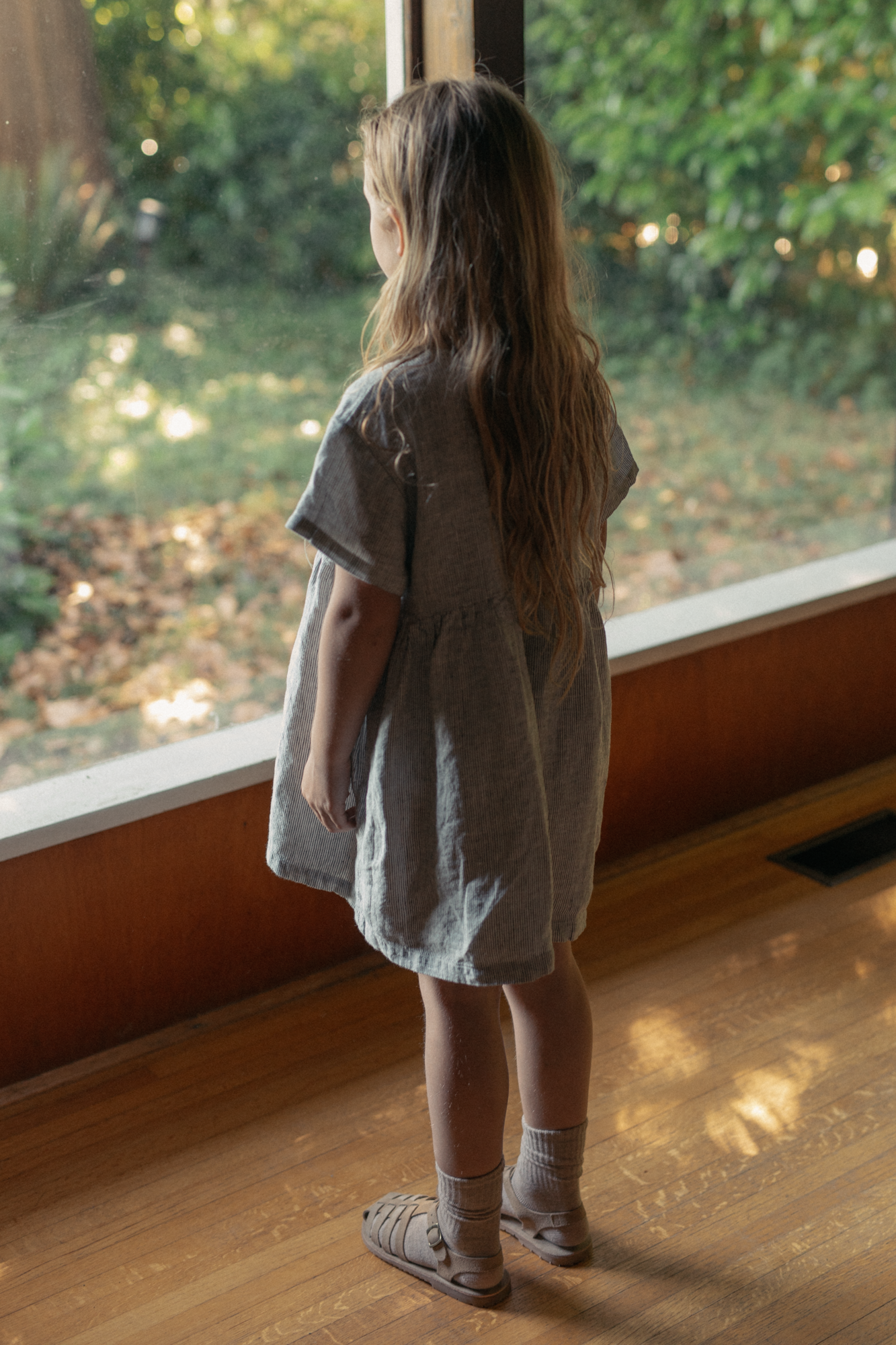 Child wearing the Mini Aya in Charcoal Stripe by Gillian Stevens, back view by a large window