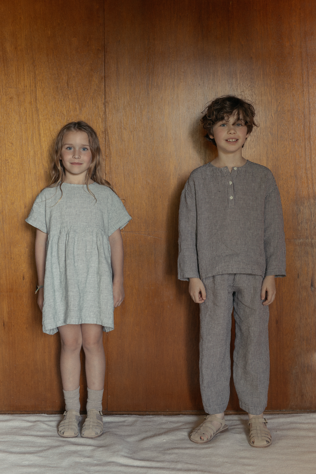 Two children wearing the Mini Aya in Charcoal Stripe by Gillian Stevens, front view against a warm wood panel