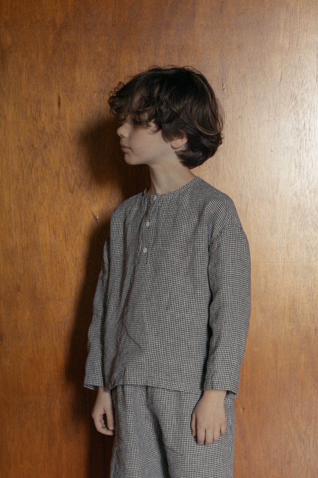 Child wearing the Bertie Top in Espresso Gingham by Gillian Stevens, side profile view against a warm timber wall