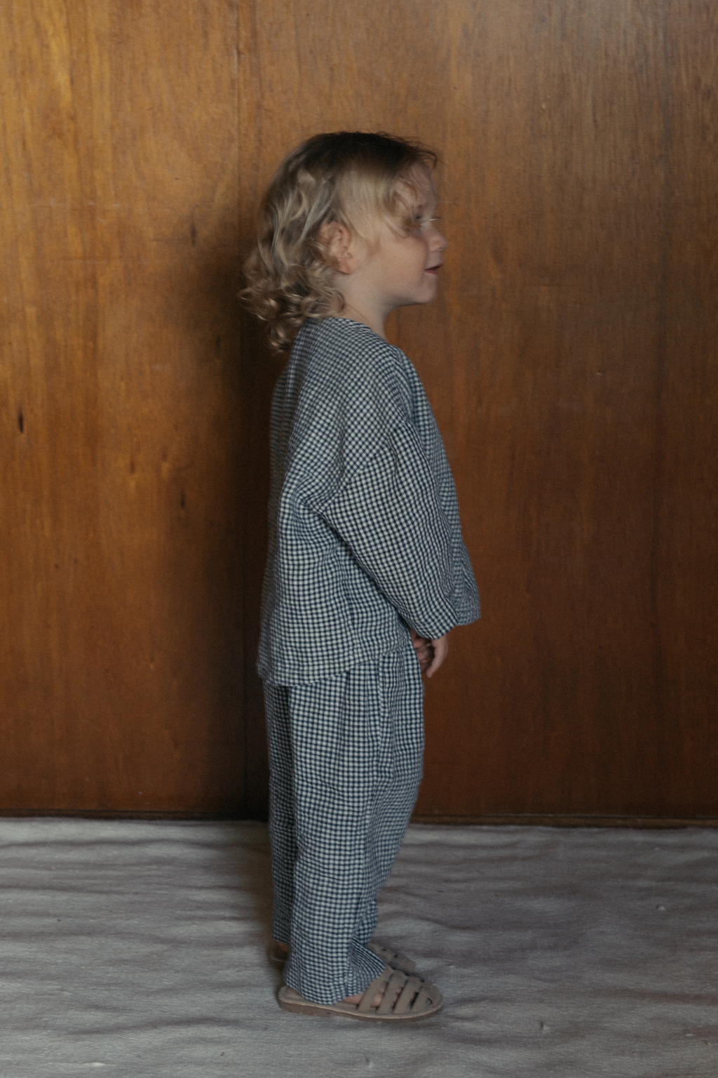 Child wearing the Bertie Top in Marine Gingham by Gillian Stevens, side view against a wood panel wall