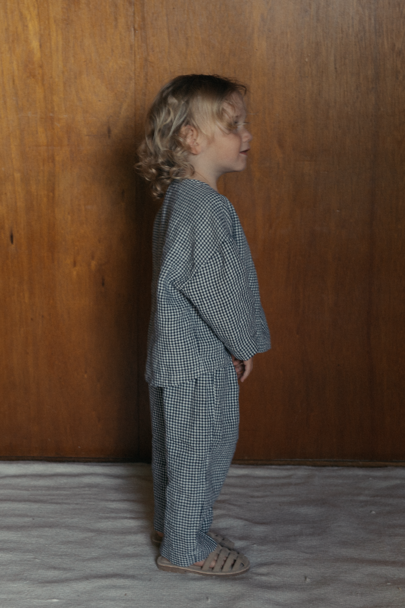 Child wearing the Bertie Top in Marine Gingham by Gillian Stevens, side view against a wood panel wall