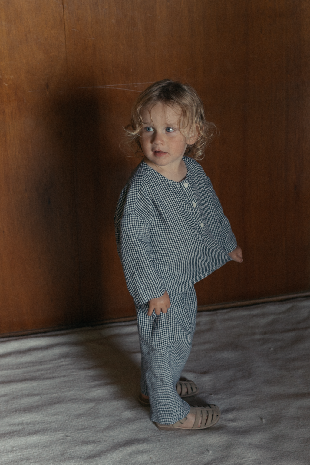 Child wearing the Bertie Top in Marine Gingham by Gillian Stevens, three-quarter view against a timber wall