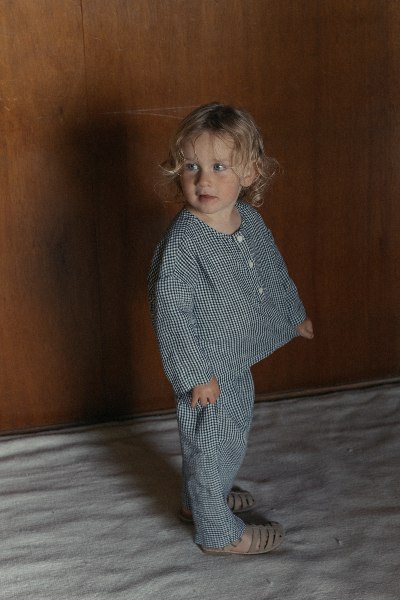 Child wearing the Bertie Top in Marine Gingham by Gillian Stevens, three-quarter view against a timber wall