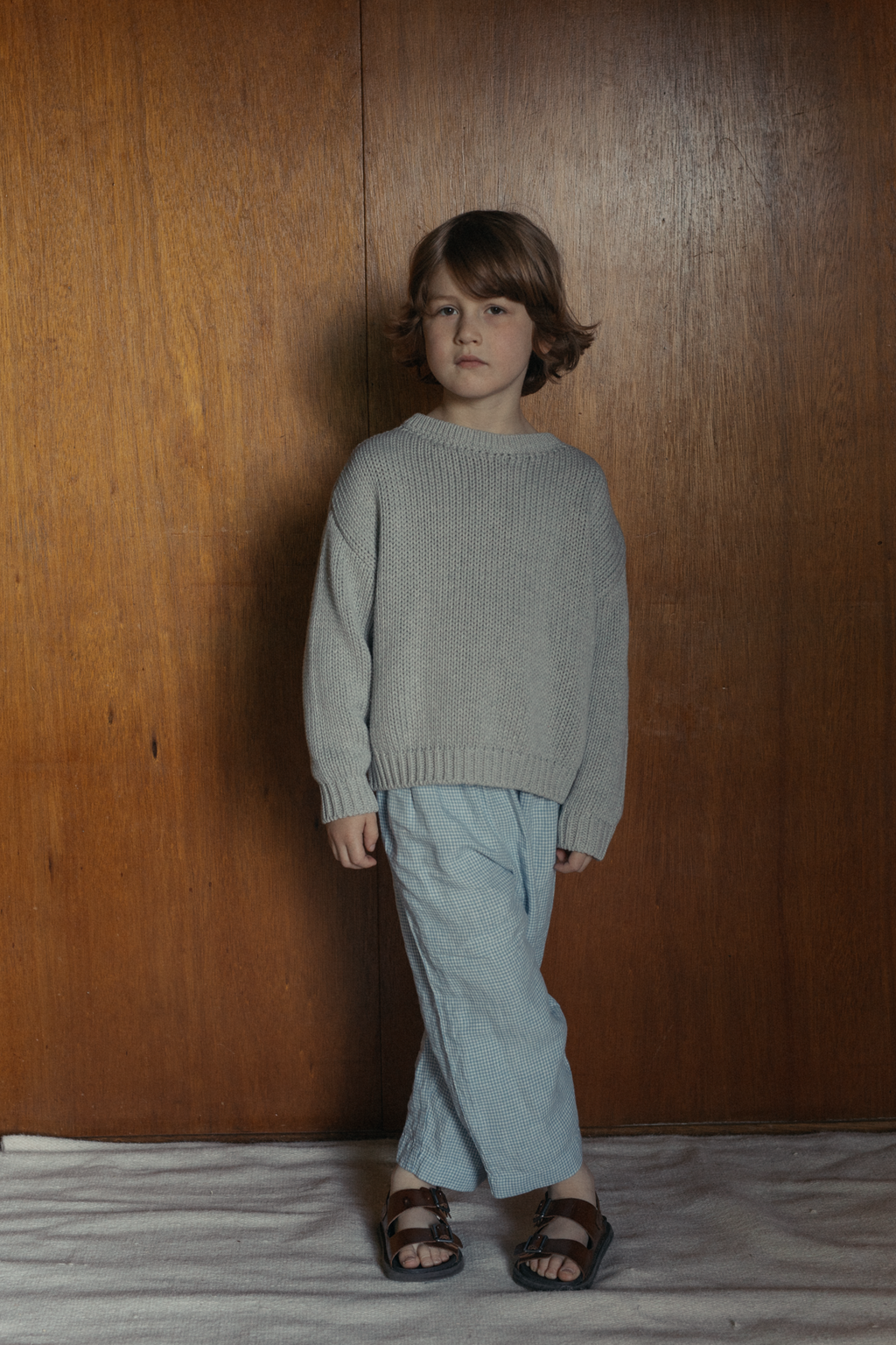 Child wearing the Mini Townes Pullover in Pebble by Gillian Stevens, front view against a warm wood panel