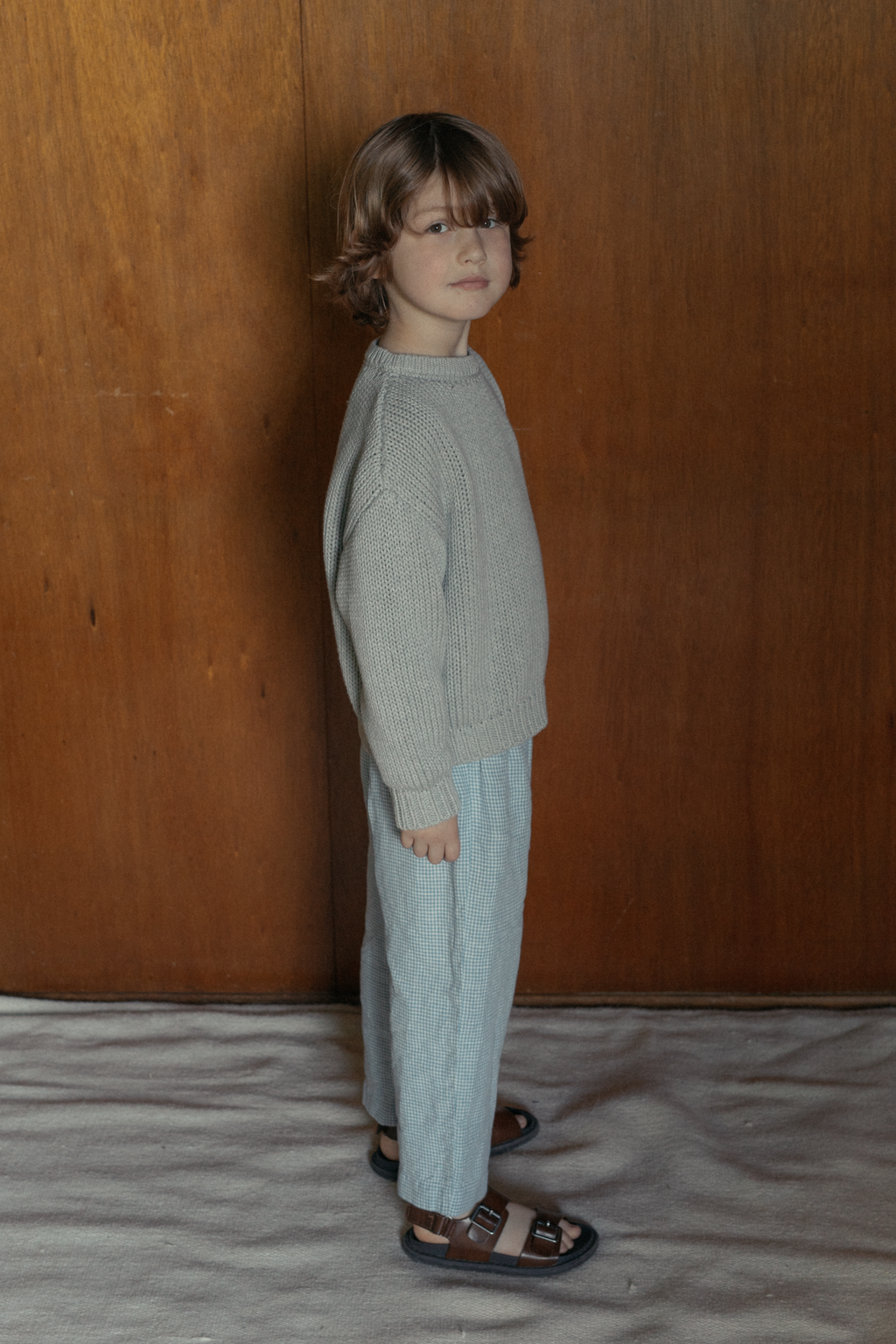 Child wearing the Mini Townes Pullover in Pebble by Gillian Stevens, side view against a wood panel backdrop