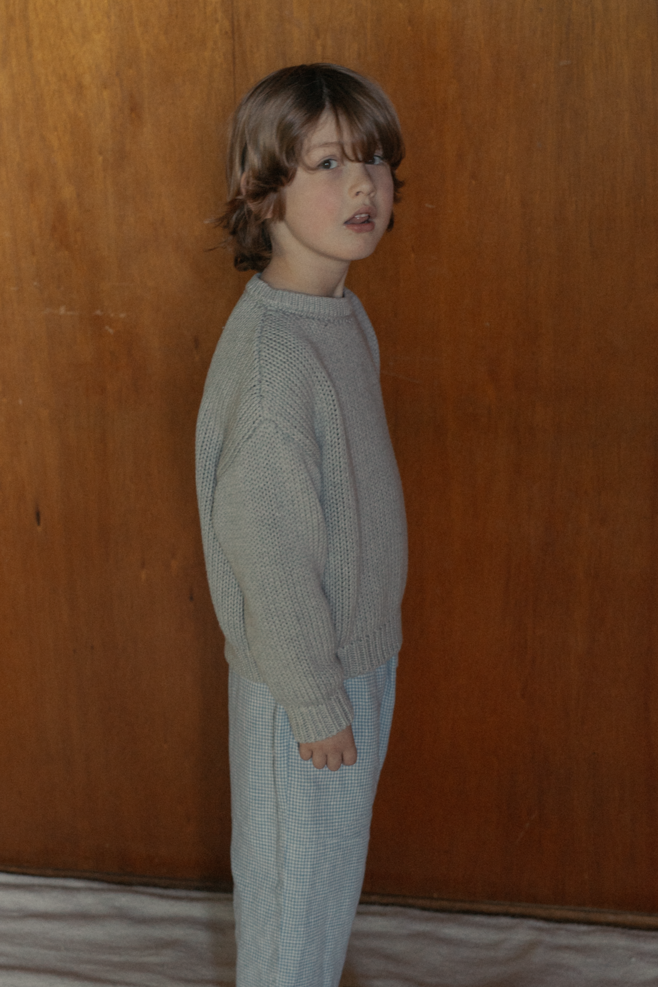 Child wearing the Mini Townes Pullover in Pebble by Gillian Stevens, side view against a wood panel background