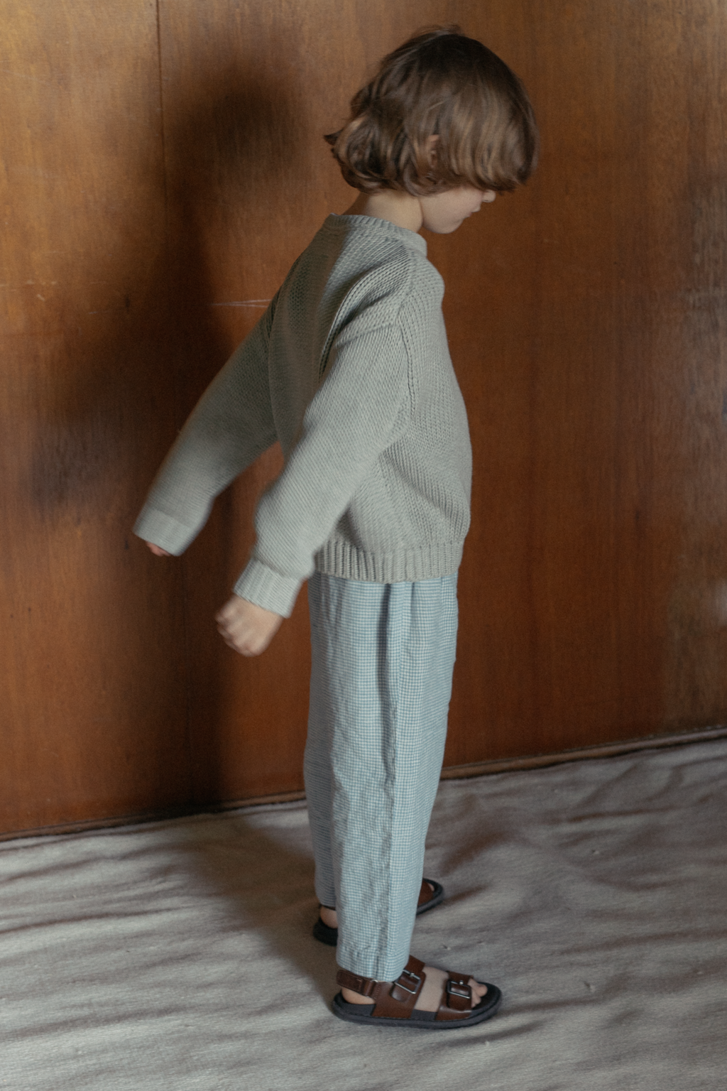 Child wearing the Mini Townes Pullover in Pebble by Gillian Stevens, side view against a wooden wall