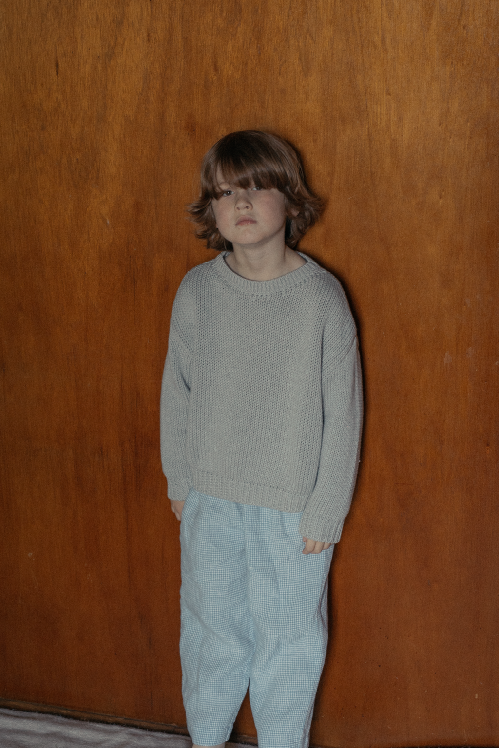Child wearing the Mini Townes Pullover in Pebble by Gillian Stevens, front view against a warm timber wall