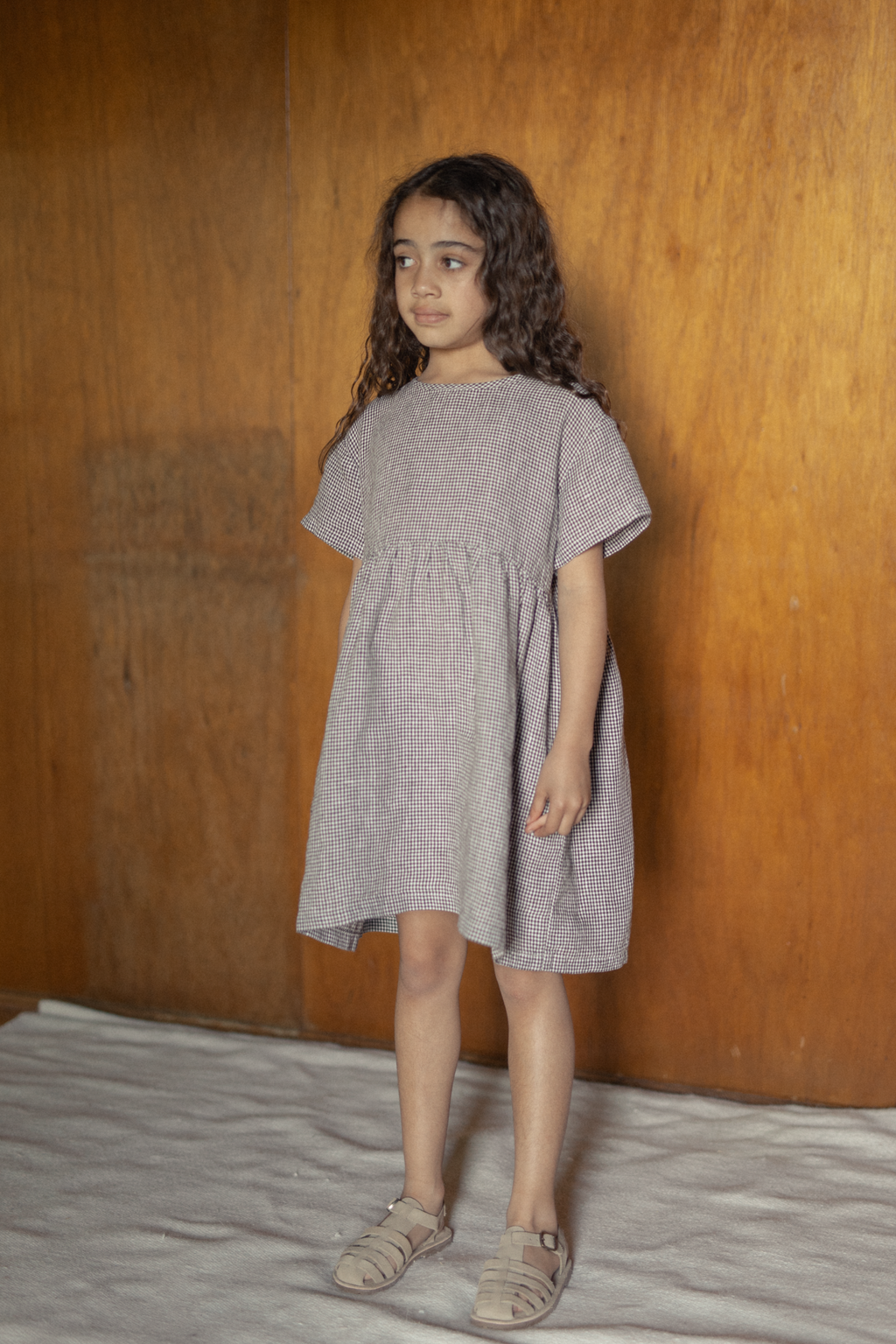 Child wearing the Mini Aya in Espresso Gingham by Gillian Stevens, front three-quarter view against a timber wall