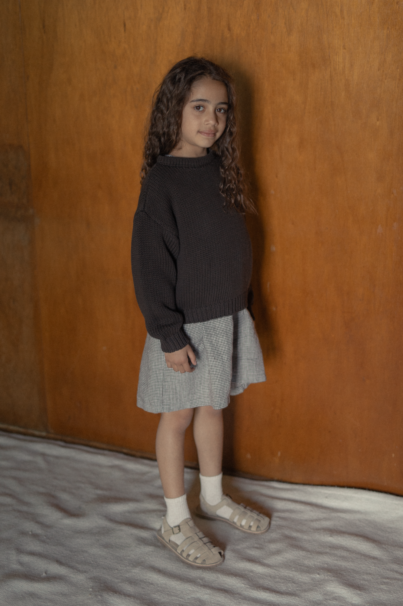 Child wearing the Mini Townes Pullover in Espresso by Gillian Stevens, three-quarter view against a warm timber wall