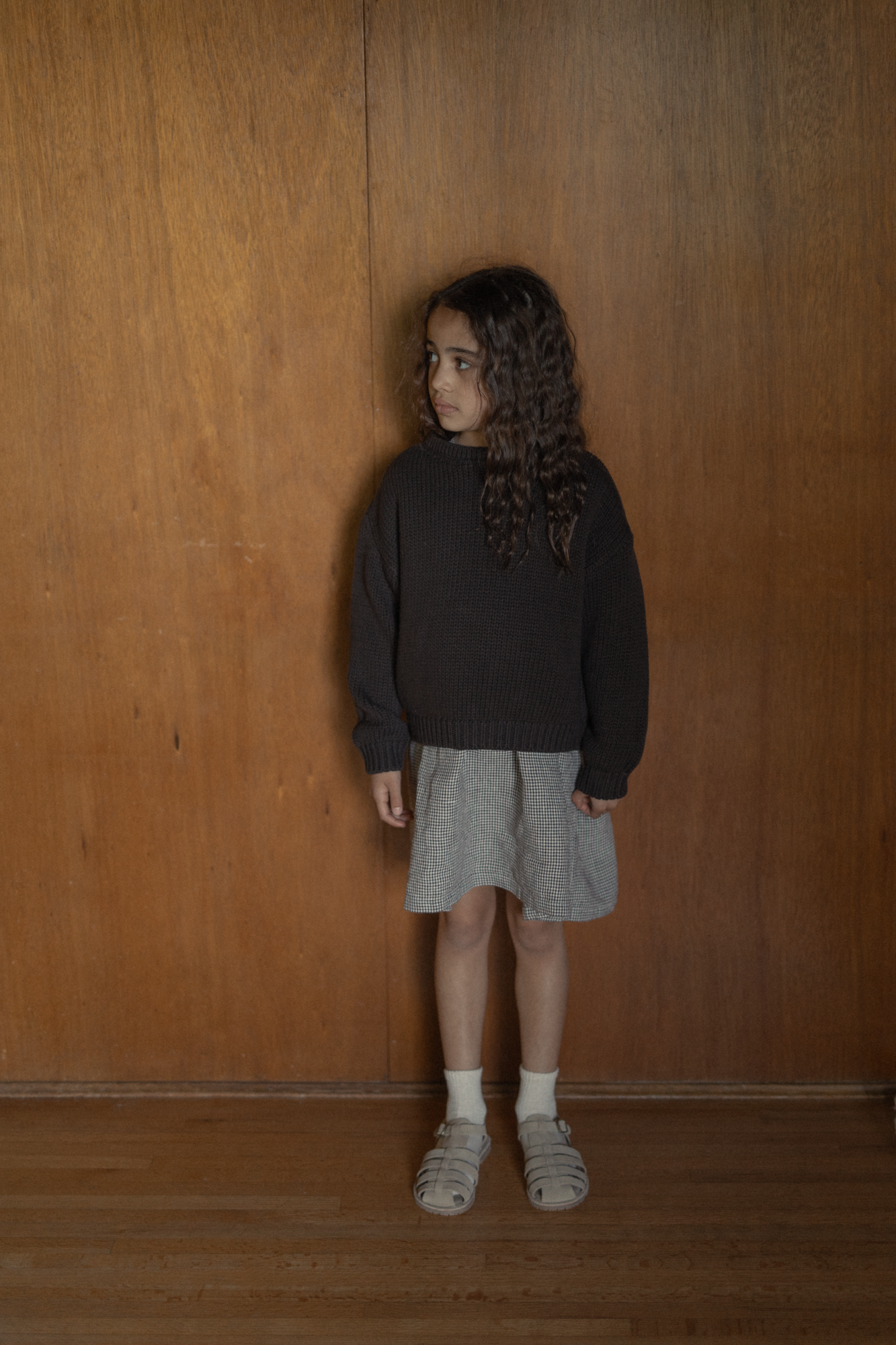 Child wearing the Mini Townes Pullover in Espresso by Gillian Stevens, front view against a warm wood-panelled wall