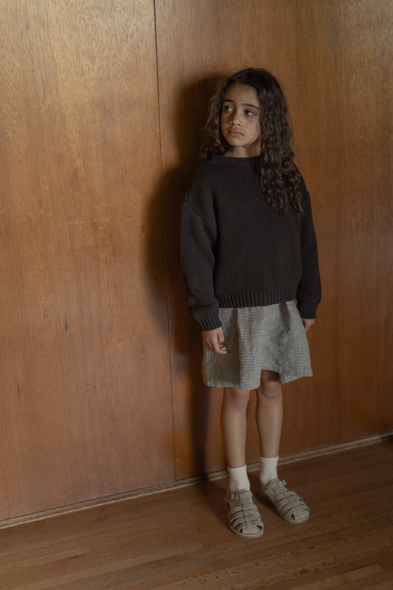 Child wearing the Mini Townes Pullover in Espresso by Gillian Stevens, full-length front view against a wood-panelled wall