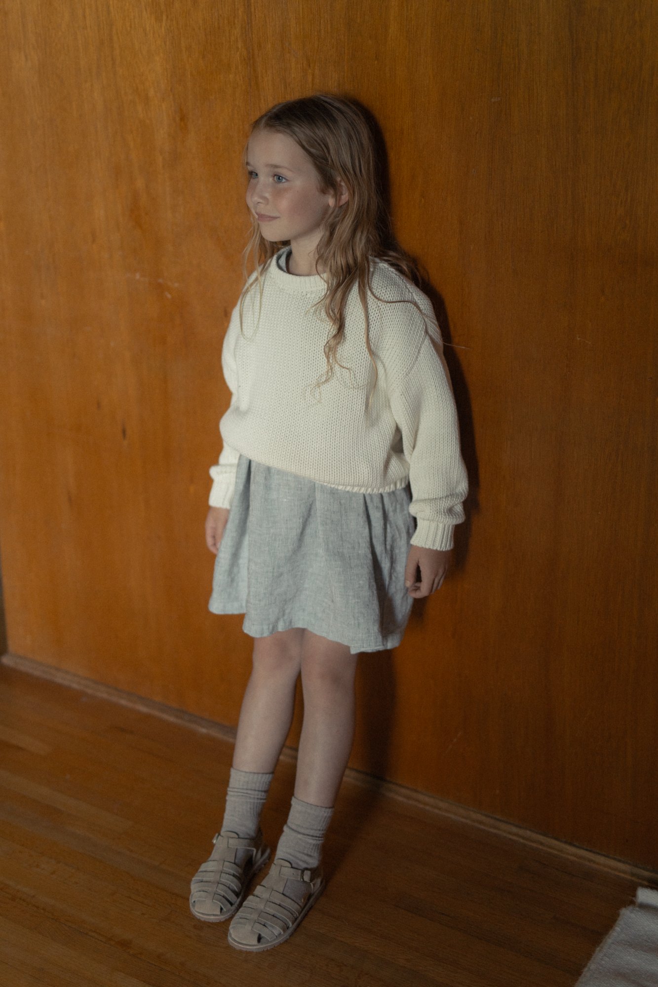 Child wearing the Mini Townes Pullover in Alabaster by Gillian Stevens, three-quarter view against warm timber wall