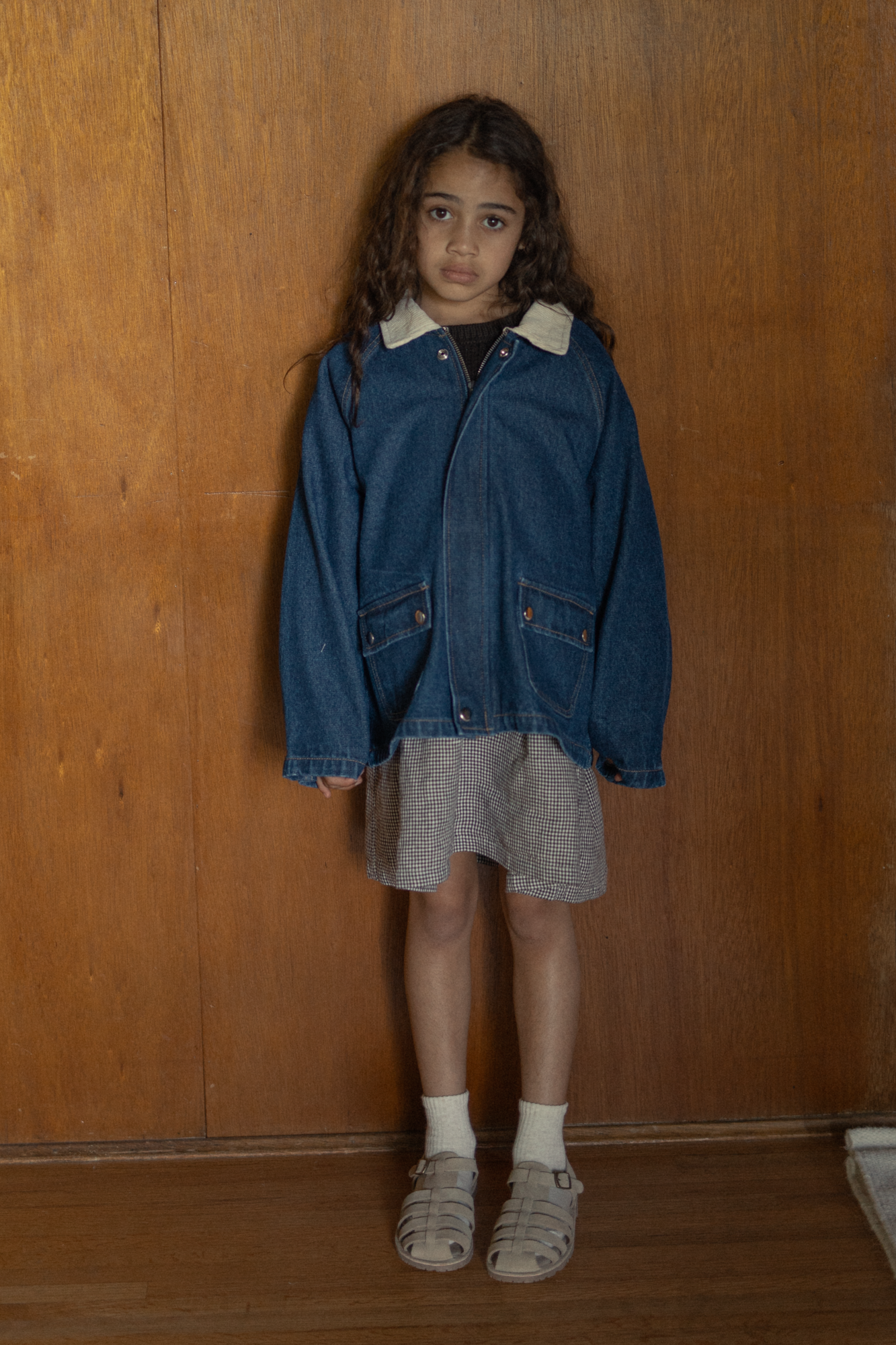 Child wearing the Brooks Jacket in Indigo by Gillian Stevens, front view styled over a check skirt against wood panelling