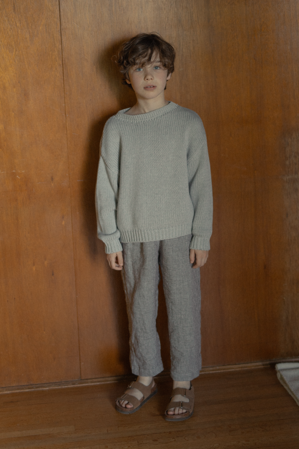 Child wearing the Mini Townes Pullover in Pebble by Gillian Stevens, full-length front view against a timber wall
