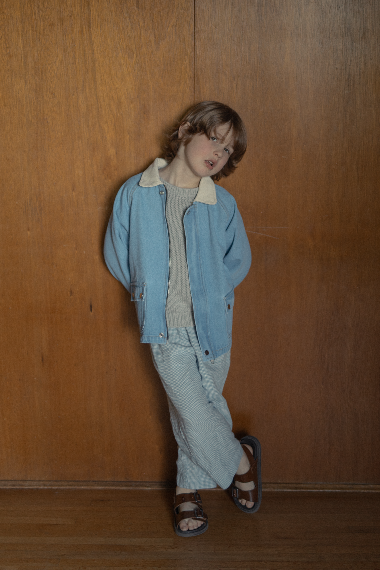 Child wearing the Brooks Jacket in Vintage Blue by Gillian Stevens, full-length front view against wood panelling
