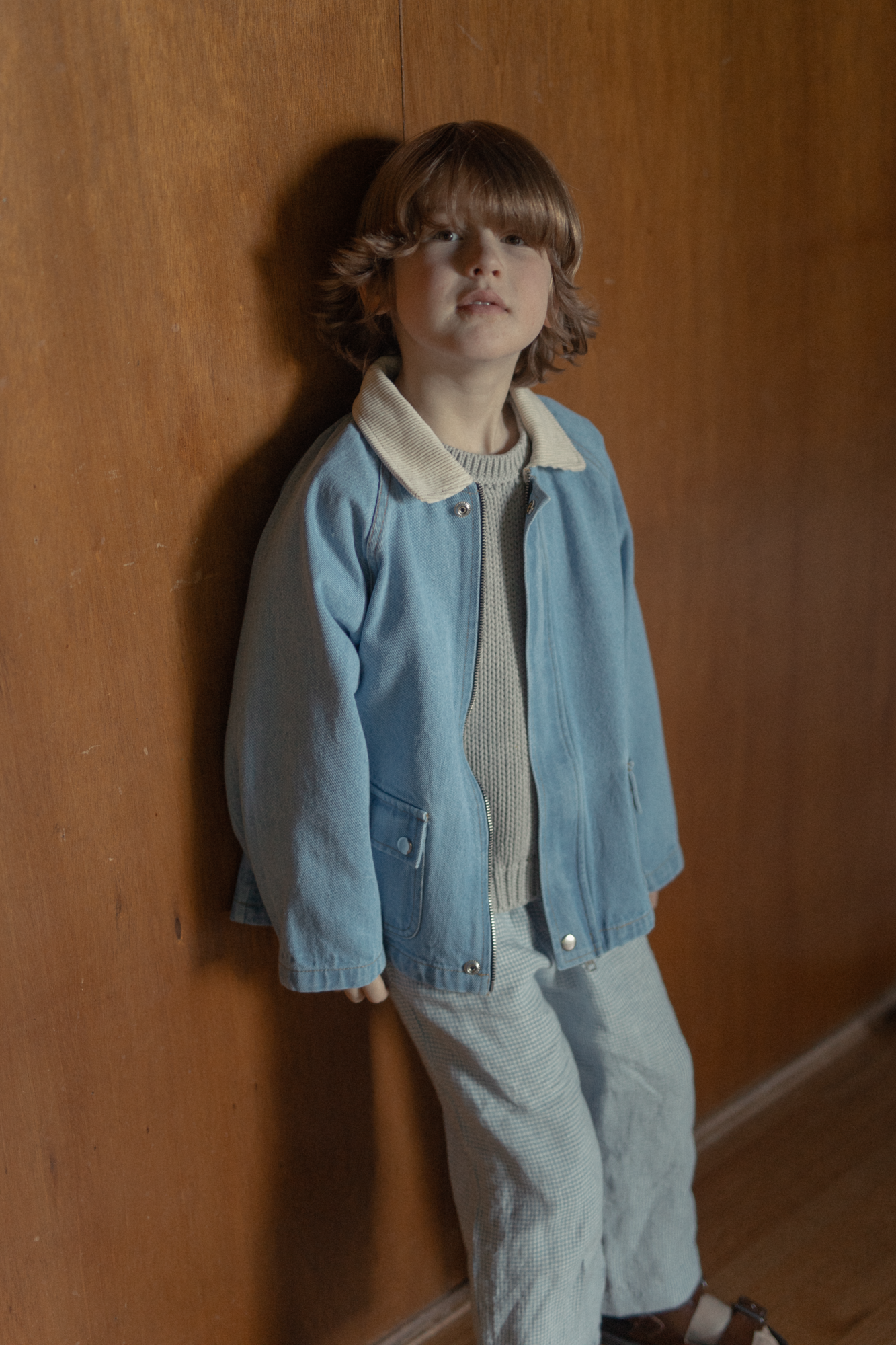 Child wearing the Brooks Jacket in Vintage Blue by Gillian Stevens, front view leaning against a wooden wall