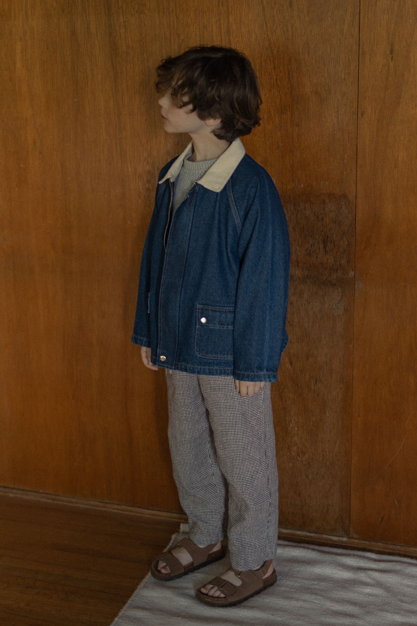 Child wearing the Brooks Jacket in Indigo by Gillian Stevens, side view against warm wooden panelling