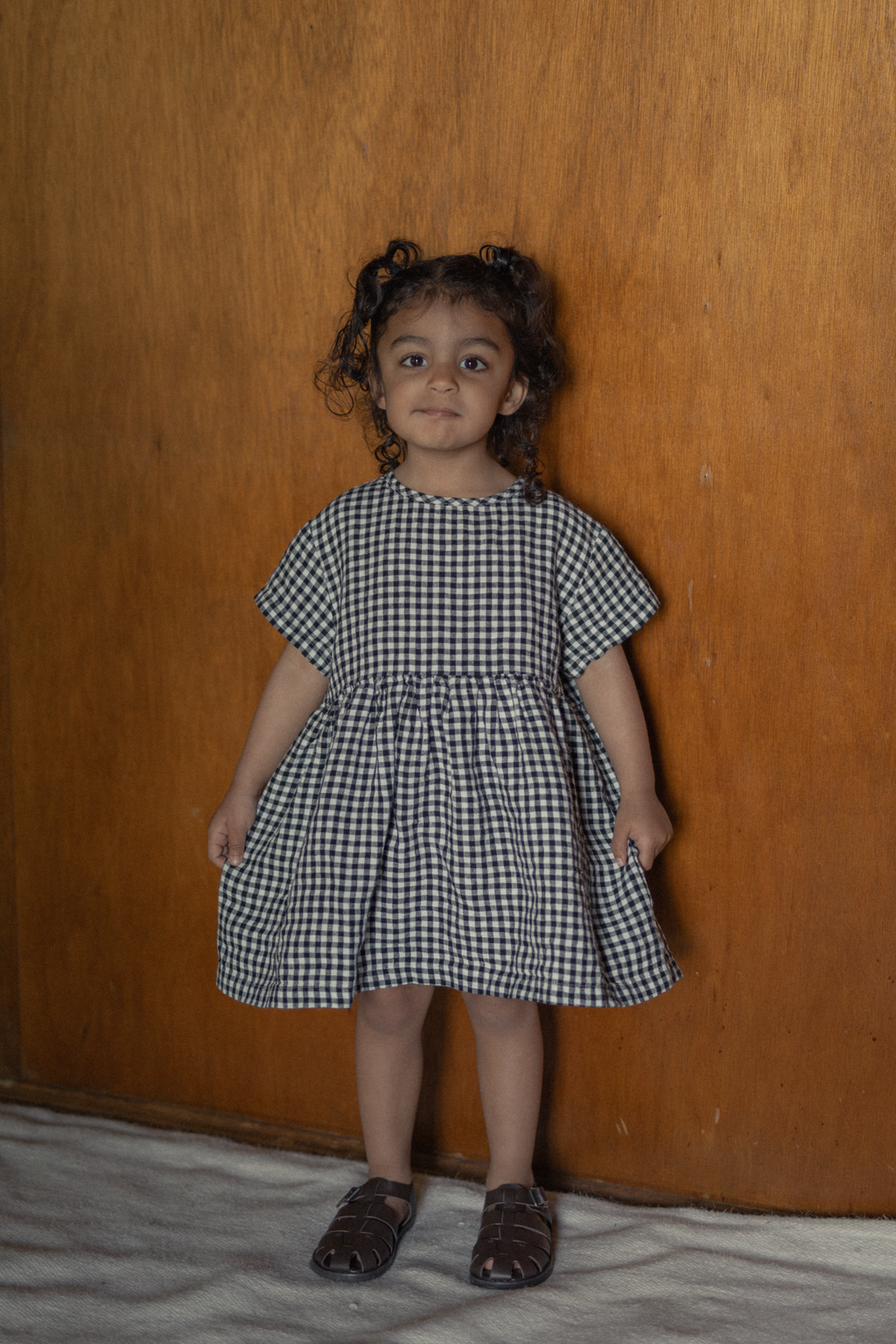 Child wearing the Mini Aya in Gingham Noir by Gillian Stevens, front view against a wood-panelled wall
