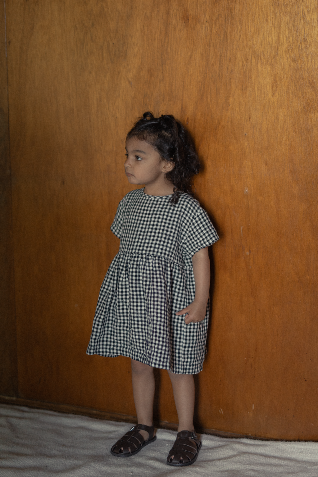 Child wearing the Mini Aya in Gingham Noir by Gillian Stevens, front view with wooden panel backdrop